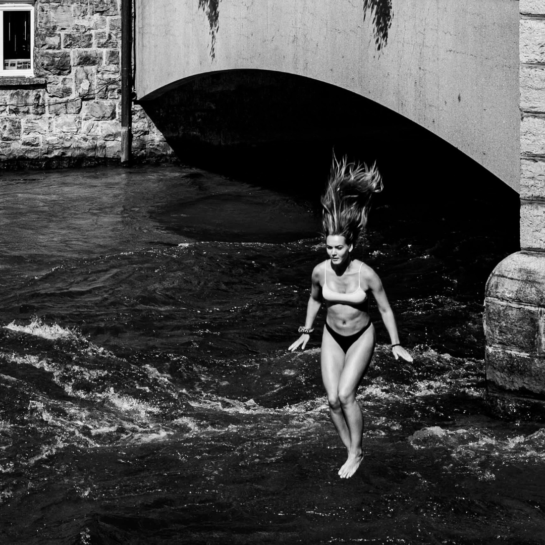 Black and white photograph of a woman emerging from a river beneath a stone bridge, dynamic movement and strong contrast, photography by Andrea Federico Santicoli.