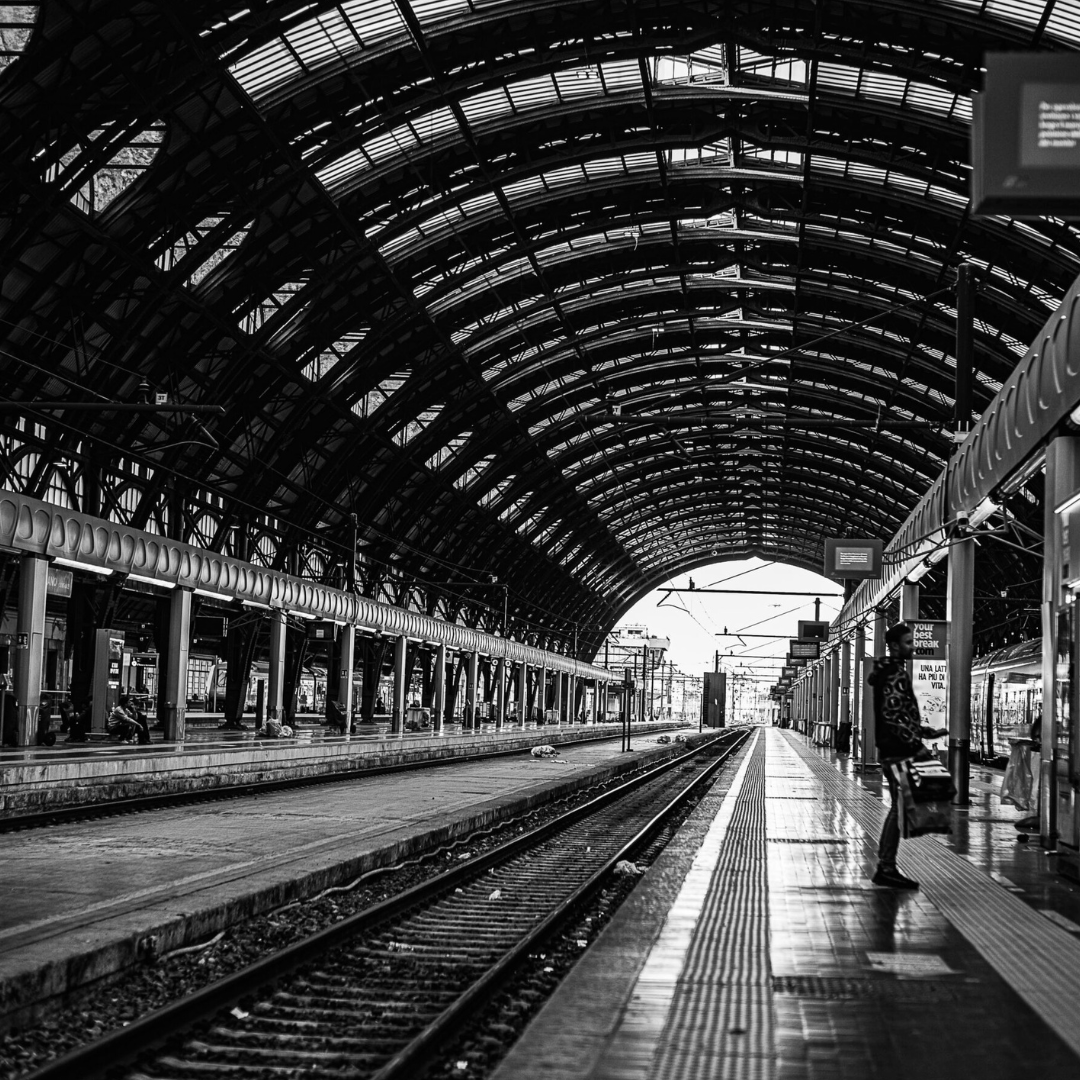 Train station platform with converging tracks and light beyond the exit, evoking arrivals and departures, black and white photography by Andrea Federico Santicoli.