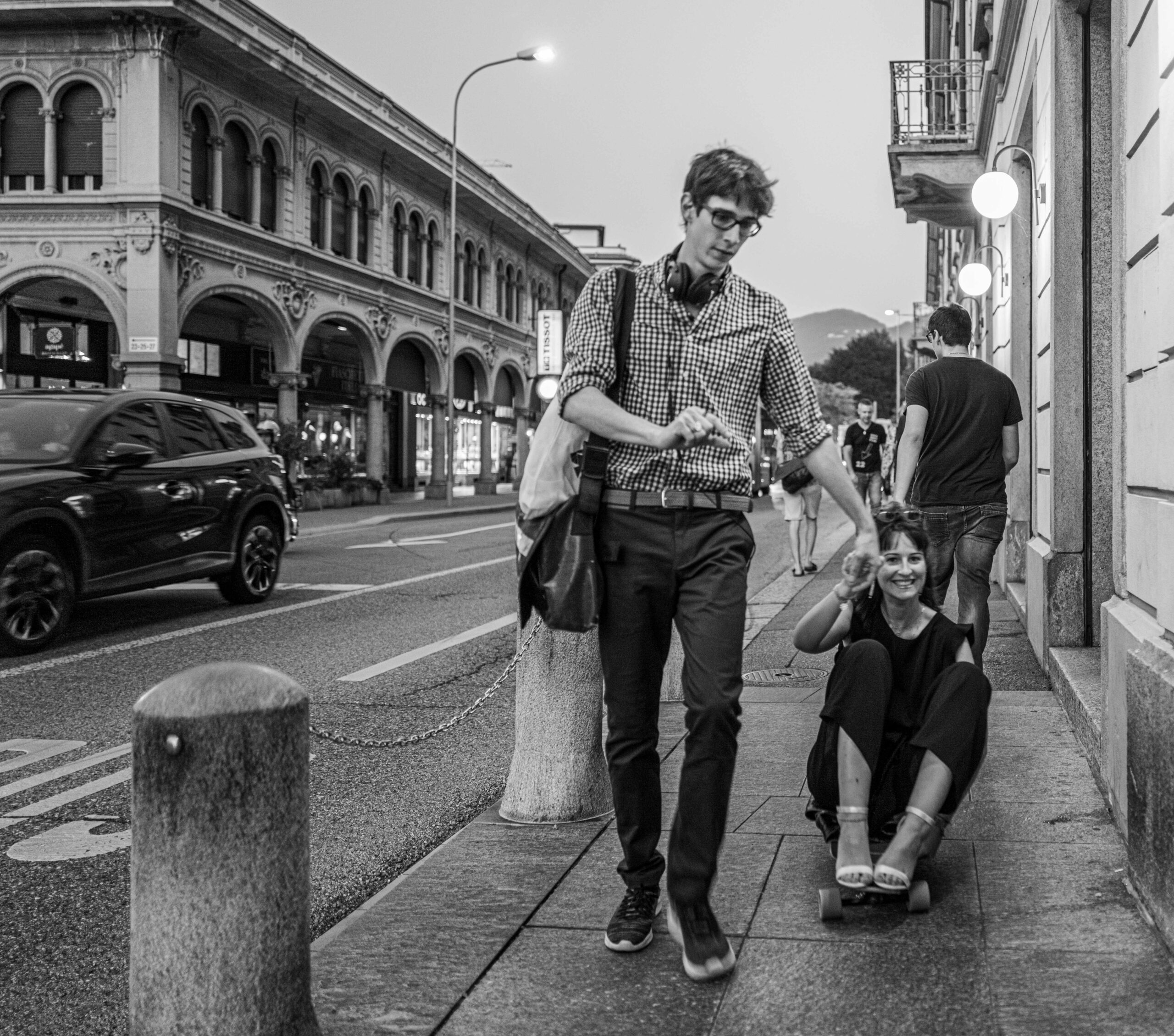 Young man pulling a smiling woman on a skateboard along a city sidewalk, black and white photography by Andrea Federico Santicoli.