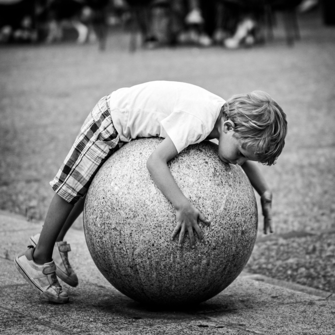 Child lying on a large stone sphere in an urban square, black and white photograph by Andrea Federico Santicoli.